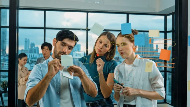 Professional business team brainstorm, sharing, thinking idea while skilled male leader write on sticky notes at glass wall. Skilled manager placed business plan on glass wall at office. Tracery