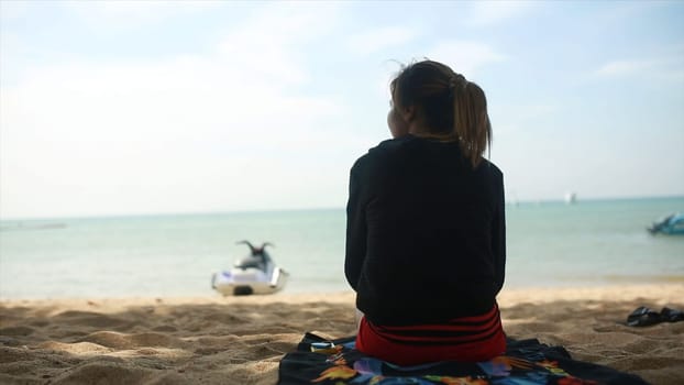 Rear view of a girl in a hoodie sitting on the beach and watch the sea, blue sky and sea background. Clip. Young woman in sportswear sitting on the beach.