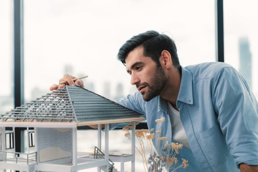 Attractive smart civil engineer measure house model roof construction. Professional architect in casual outfit looking at house model while planing building structure. Skyscraper view. Tracery.