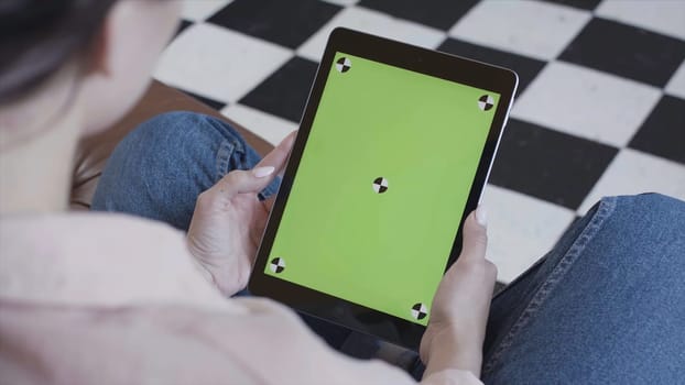 Young brunette woman watching her tablet with green screen on black and white floor background. Side view of a girl using tablet pc with chroma key.