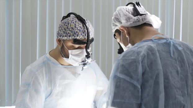 Two surgeons working at the operating clean room. Two male doctors wearing sterile uniform and binocular loupe while performing surgery, concept of health care and medicine.