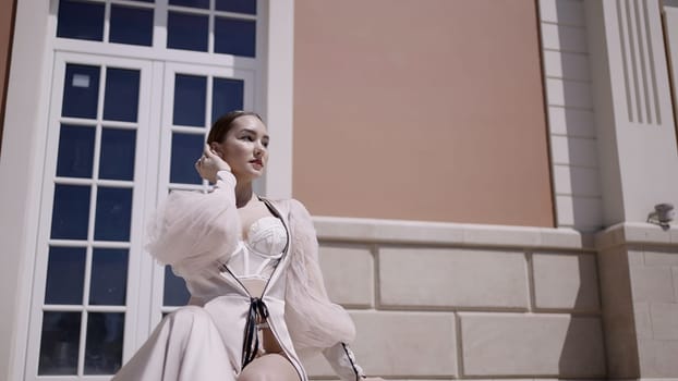 Beautiful model in white lingerie and cape. Action. Young woman sitting next to red buildings and posing for a photographer. High quality 4k footage
