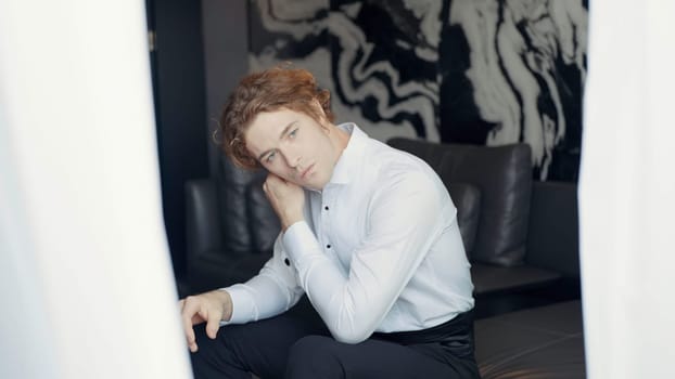 A man with a white dress shirt. Action. A man with red hair is sitting on the sofa and adjusting the collar of his clothes against a background of light black wallpaper and a bright white curtain in the foreground