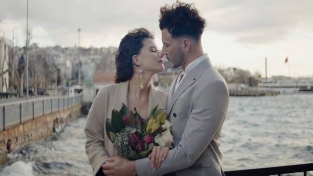 Beautiful wedding photo shoot with two models.Action.A young beautiful couple with a tall groom in suits and a bouquet of flowers are standing on the embankment and flirting with each other. High quality 4k footage