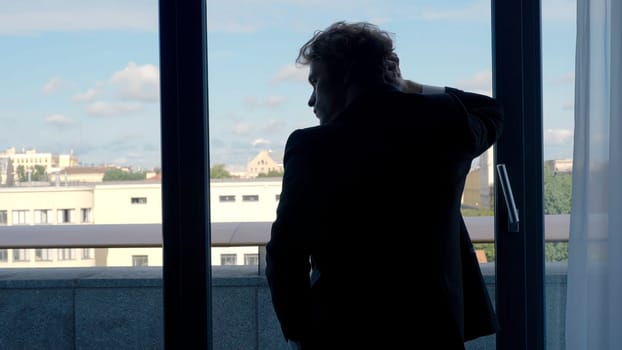 A man in a suit with long hair.Action. A handsome man stands at the window and looks at the city holding his hand behind his head, the sky is blue and small city houses are on the street