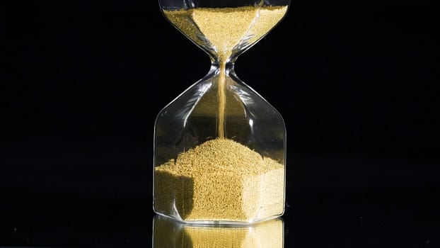 Close-up of golden hourglass. Stock footage. Stylish hexagon hourglass with falling gold specks on black isolated background. Passage of time and importance of every grain.
