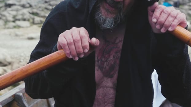 Old man rowing oars in wooden boat. Stock. Old man with beard and tattoo on chest quickly sail from shore on wooden boat alone. Close-up of old man strongly rowing oars.