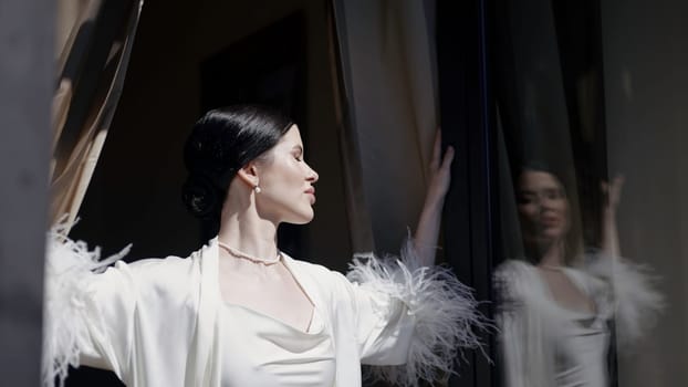 Posing brunette in a sexy white cape. Action. A beautiful woman posing in profile with an open window and the sun. High quality 4k footage