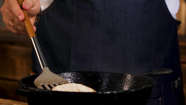 Close up of male cook frying camembert cheese in a black iron pan from the both sides with a metal shovel. Process of food preparation at the restaurant.