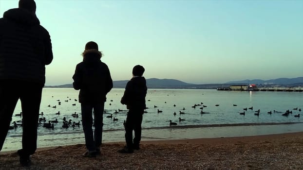 Father and two sons throwing stones into the water on the shore of the sea. Father and little son, silhouettes on beach at sunset.