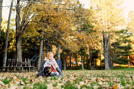 Little girl sits on the belly of her mother lying on the green grass among the fallen leaves. High quality photo