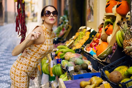 Side view of young stylish female model in trendy armless gown and eyeglasses standing on street and leaning towards stall with assorted fruits on street