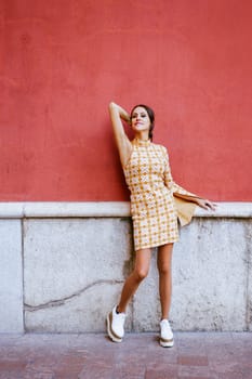 Full body of young sensual female model in makeup with stylish dress and trendy footwear standing on street raising elbow while looking at camera