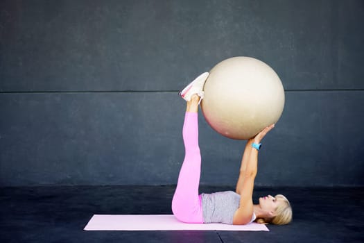 Side view of fit young female in activewear lying on mat, and doing exercise with fitness ball while practicing workout against gray wall at modern gym