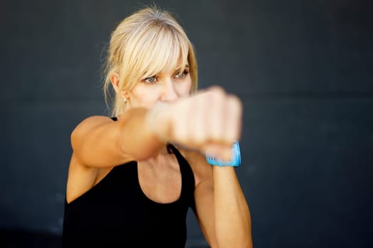 Serious young fit female boxer in black sportswear practicing punches during boxing training against gray background looking away pensively while workout at gym