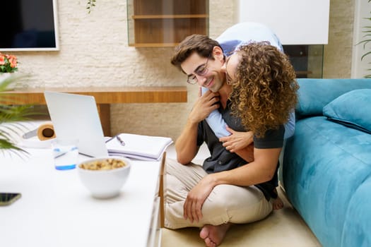 Loving young woman embracing man from behind while sitting near laptop and notebook in living room at home