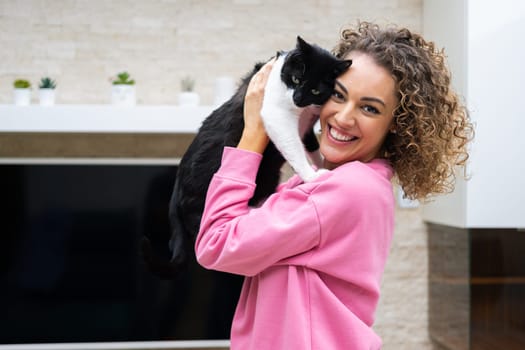 Side view of cheerful female with curly hair in casual cloth smiling and looking at camera while embracing cute cat in bright room