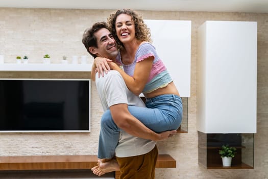 Side view of young cheerful couple in casual clothes hugging and standing at home while enjoying time together against TV with potted plants on shelves