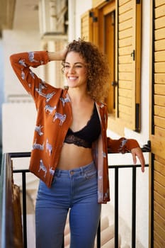 Young slim woman wearing stylish outfit leaning on balcony railing while looking away in modern apartment