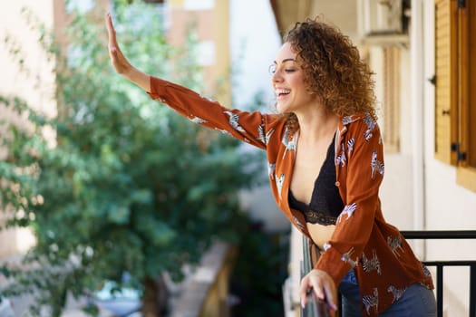 Side view of smiling curly haired female in stylish clothes standing on balcony and greeting with raised hand while looking down against blurred background
