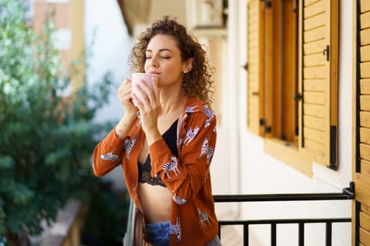 Young curly haired woman enjoying coffee while leaning on balcony of modern building against blurred background in daytime