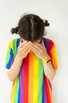 This is a portrait of a girl dressed in a shirt with a rainbow covering their face with her hands. The background is white. The mood of the image is sad or upset