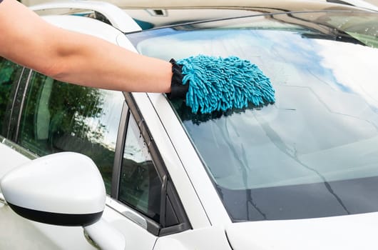 A person s hand with a blue microfiber mitt cleaning the windshield of a white car