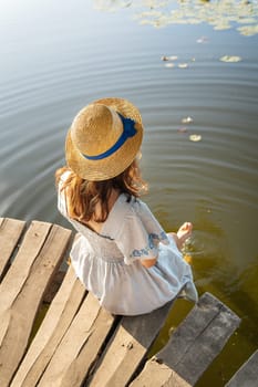 A young beautiful girl in a hat and a long dress sits with her back on the pier by the lake. Outdoor recreation concept