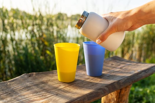 A hand, pouring tea from a white thermos into a blue cup on a wooden table with a yellow cup next to it. The background is a lake with reeds