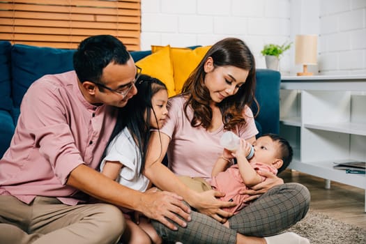 A loving mother sits in the living room feeding her newborn baby with a milk bottle while her toddler son and daughter bond with dad. Family togetherness is the heart of joy at home.