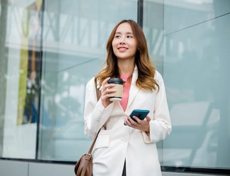 Young business woman smiling holding mobile phone with coffee take away going to work early in morning, Asian businesswoman with smartphone and cup coffee standing against street building near office