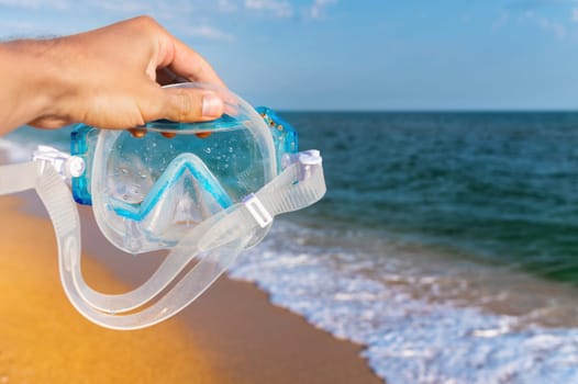 An outstretched male hand holds a snorkeling mask on the sea of sea waves and a golden sandy beach. necessary equipment for underwater sports.