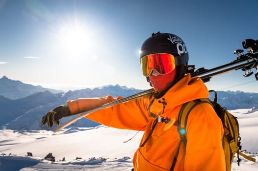 Portrait of a confident skier in an orange jacket standing on the top of a mountain and holding skis in his hands. Happy man enjoying ski holiday on sunny winter day.