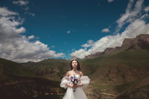 The bride with a wedding bouquet, her eyes closed and her face basking in the sun in the mountains. panoramic view of mountain range from behind of married young woman.