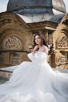 A bride in a white dress sits on a ledge with an old building in the background. Young Caucasian woman straightens her hair at a wedding in an ancient city.