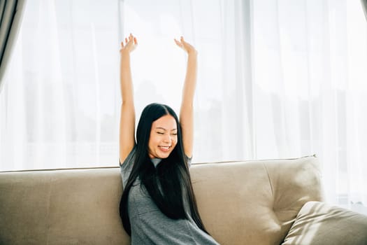 Smiling woman in luxury living room raises arms stretches after sitting. Lifestyle of a cheerful entrepreneur on sofa. Wellbeing joy and carefree relaxation.