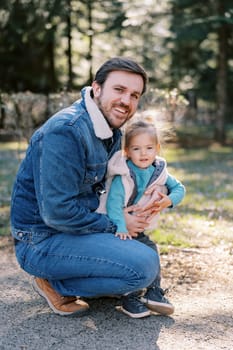 Smiling dad squatting and hugging little daughter in park. High quality photo