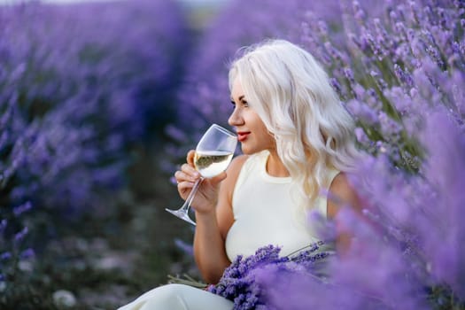 Blonde lavender field holds a glass of white wine in her hands. Happy woman in white dress enjoys lavender field picnic holding a large bouquet of lavender in her hands . Illustrating woman's picnic in a lavender field