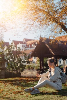 Young fashionable teenage girl with smartphone in park in autumn sitting at smiling. Trendy young woman in fall in park texting. Retouched, vibrant colors. Beautiful blonde teenage girl wearing casual modern autumn outfit sitting in park in autumn. Retouched, vibrant colors, brownish tones.