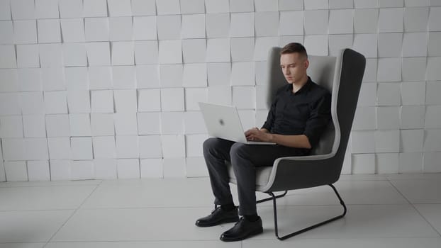 Young successful man in chair with laptop. Action. Young guy is working on laptop in business atmosphere. Business style chairs and rooms with successful man.