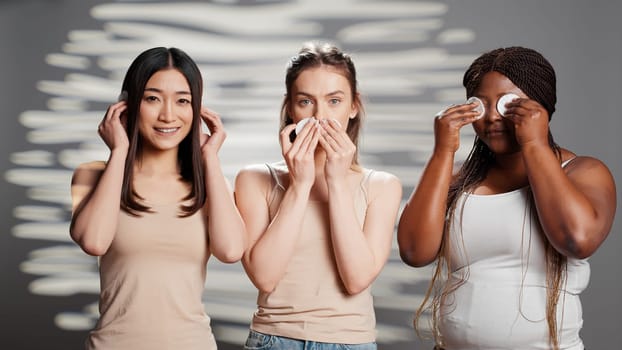 Young models doing three wise monkeys sign with cotton pads, creating beauty skincare campaign in studio. Diverse women having fun playing around with cosmetics, cleansing skin.