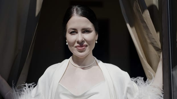 Happy smiling woman in white clothes in a hotel room. Action. Brunette woman against the sunlight wearing a robe with feathers