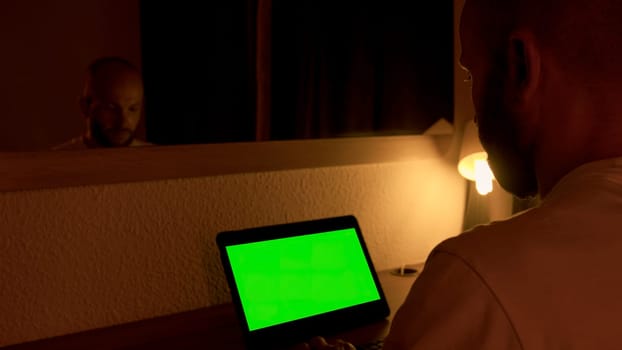 Man sitting at his desk, works on a laptop with green chroma key screen. Media. Man uses portable computer for work at home at night in front of mirror