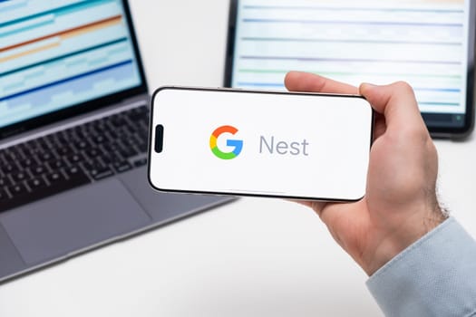 Nest logo on the screen of a smartphone in mans hand with laptop and tablet with graphical diagrams on the background, December 2023, Prague, Czech Republic.