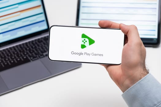Google Play Games logo on the screen of a smartphone in mans hand with laptop and tablet with graphical diagrams on the background, December 2023, Prague, Czech Republic.