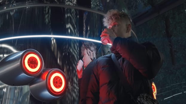 Young man at futuristic interior with lights. Action. Man drinks drink from cup at mirrored wall. Man stands by mirrored futuristic column with red lanterns.