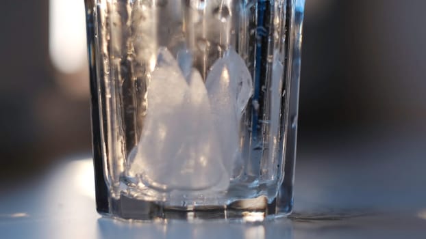 Wet empty transparent glass with ice pieces. Close up of putting ice pieces into the faceted glass standing on white table on blurred background.