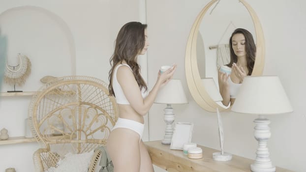 Woman stands at mirror and takes care of herself. Action. Sexy woman performs care in bright interior. Attractive well-groomed woman applies cream.