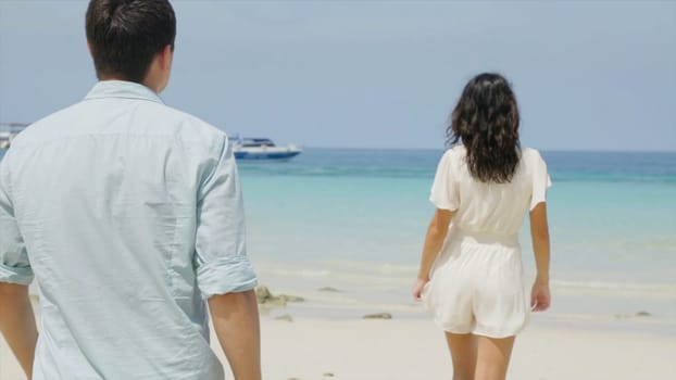 Cute couple in white clothing goes hand beach. Couple of teenagers running and flirting on the beach shore near the water. Woman walking on romantic honeymoon beach holidays holding hand of boyfriend following her, view from behind. A young girl holding hands on the beach.