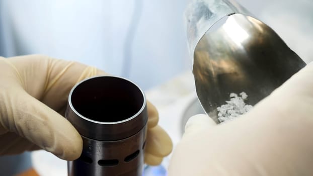 Close up of hands in medical gloves putting chemical powder from chemical scoop to small metal container at laboratory. Hands putting white reagent into chemical receptacle, science conceept